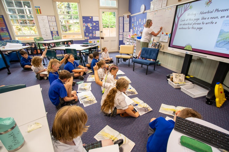 Students sitting on floor with work books while teacher is writing on whiteboard