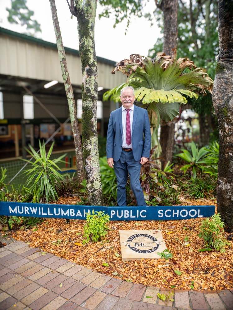 Principal standing in front of school sign and 150 year commemoration sandstone tile